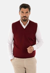 Maglione senza maniche a V in misto lana bordeaux, indossato sopra una camicia bianca con bottoni. Pantaloni beige completano l'outfit.