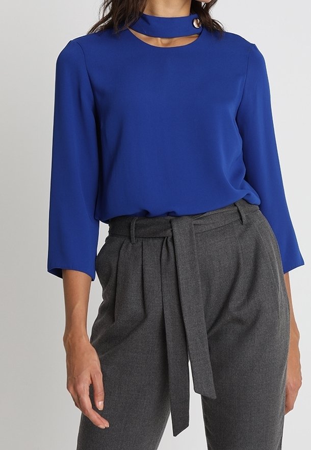 Femme portant un haut bleu à manches longues avec un col montant et un détail œillet, associé à un pantalon gris taille haute avec une ceinture en tissu.