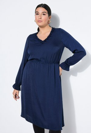 Kvinde iført langærmet navy blå kjole med flæset V-udskæring og elastisk talje, stående mod hvid baggrund med hånd på hoften.