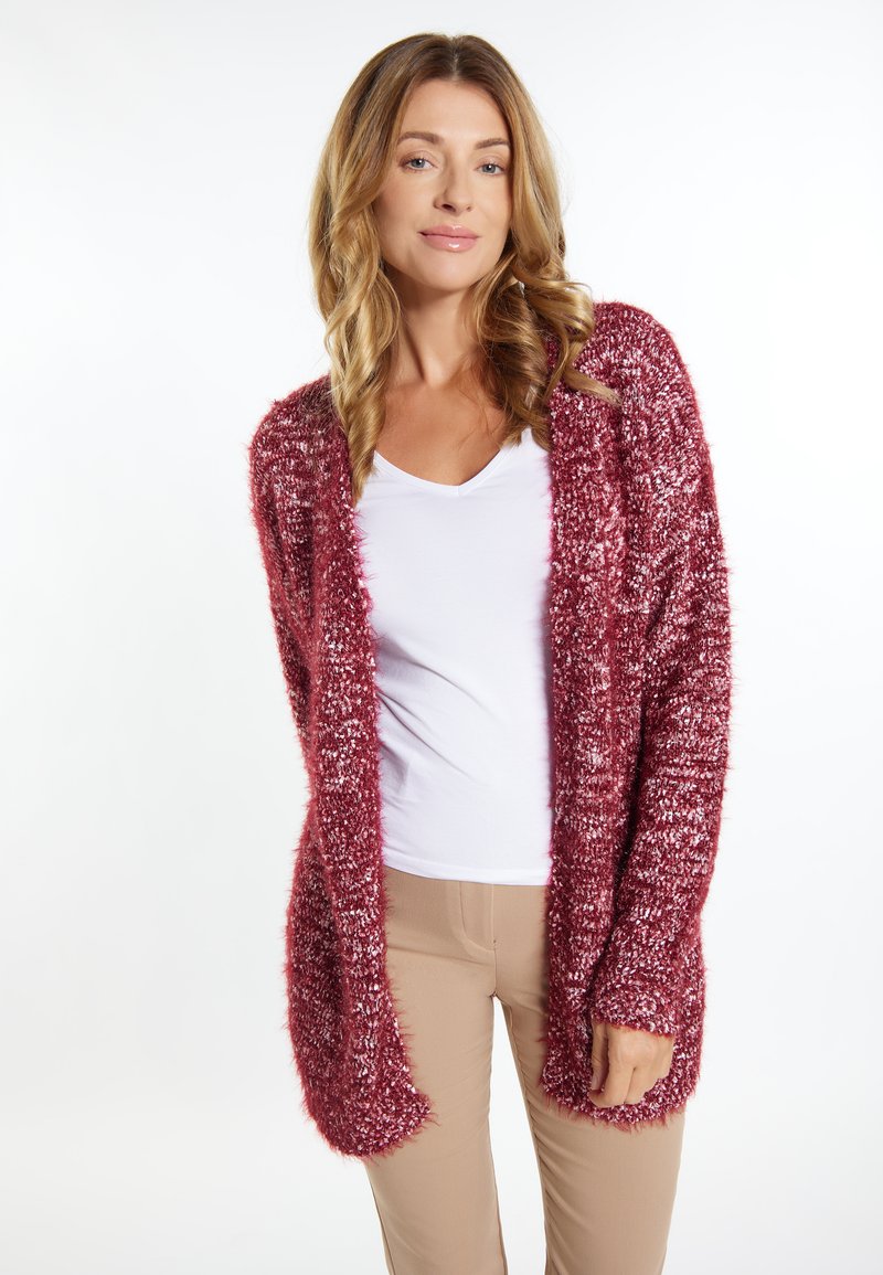 Cardigan lung burgund cu o textură pufoasă și o croială lejeră, având un front deschis și manșete cu nervuri, purtat peste un top alb și pantaloni bej.