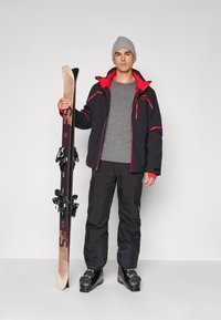 Un model de echipament de schi îmbrăcat cu o jachetă neagră cu accente roșii, cămașă gri, pantaloni negri și bocanci de schi, ținând în mână schiuri negre cu legături.
