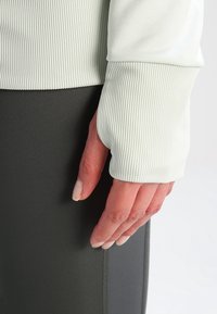 Main reposant contre des leggings noirs, portant un haut blanc côtelé à manches longues avec un poignet prolongé couvrant une partie de la main.