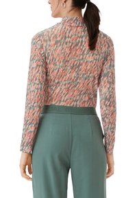 Langarmshirt mit einem mehrfarbigen, abstrakten Muster in Koralle- und Grautönen, kombiniert mit hochgeschnittenen, grünen maßgeschneiderten Hosen.