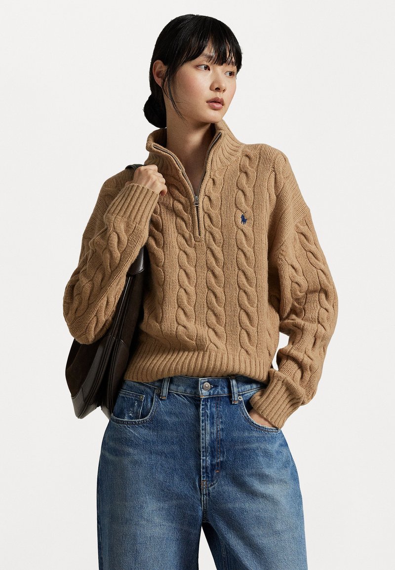 Maglione beige a trama cable knit con colletto alto e zip. Maniche e orlo a costine. Abbinato a jeans denim blu. Modella che tiene una borsa scura.
