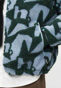 Ανοιχτό μπλε ζακέτα φλις με σκούρα πράσινα αφηρημένα υφασμάτινα σχέδια και ορατό φερμουάρ, φορεμένη πάνω από λευκό πουκάμισο και καφέ παντελόνι.