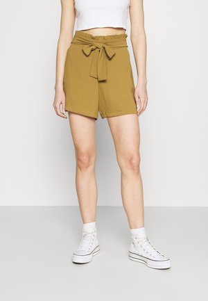 Femme portant un short taille haute moutarde avec une ceinture nouée, un haut court blanc, des chaussettes blanches et des baskets montantes blanches, debout sur un sol uni.
