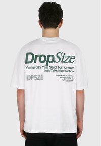 T-shirt en coton blanc à coupe décontractée. Le texte au dos comprend "Taille de Drop", "Hier tu as dit demain", et d'autres détails en vert.