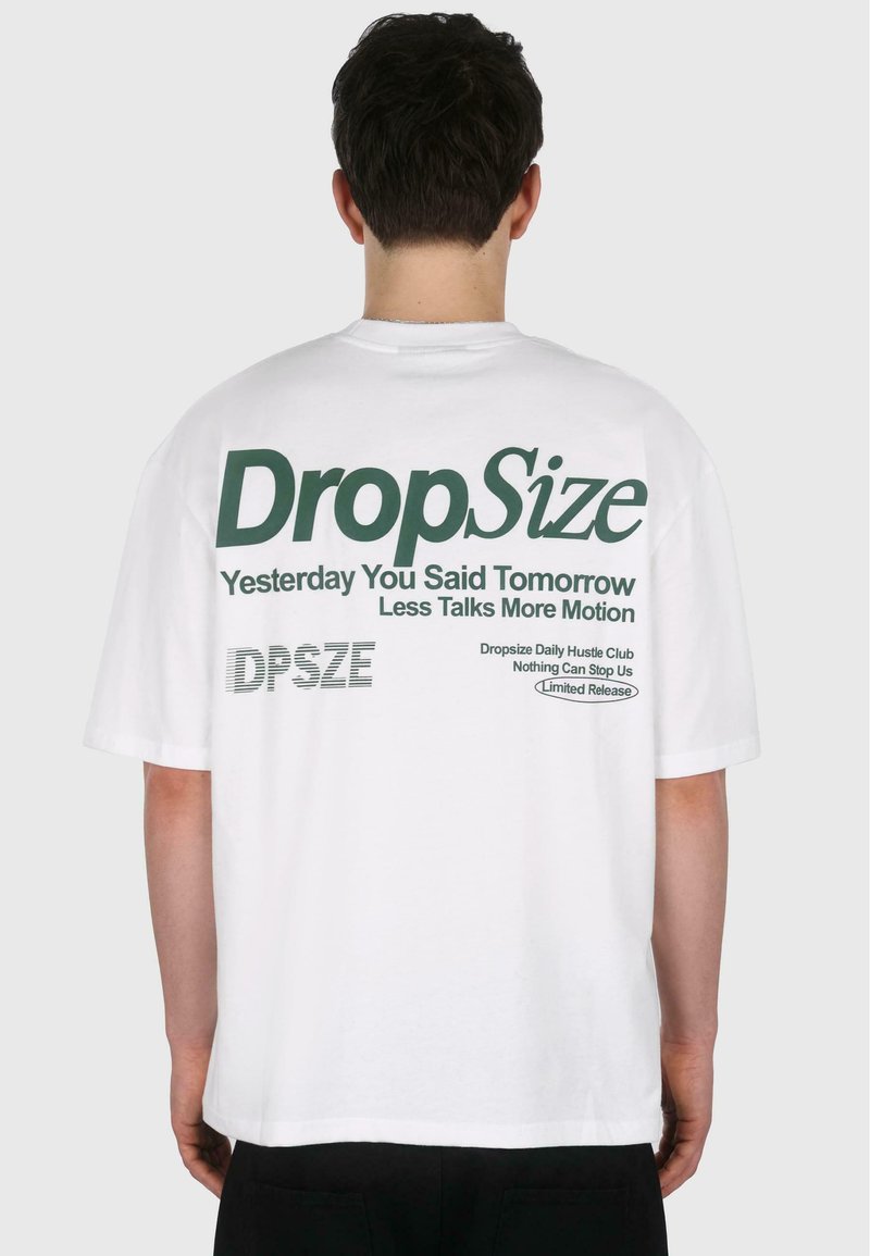 T-shirt en coton blanc à coupe décontractée. Le texte au dos comprend "Taille de Drop", "Hier tu as dit demain", et d'autres détails en vert.