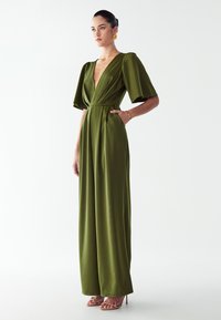 Ženska, oblečena v olivno zeleni satenski kombinezon z globokim V-izrezom, širokimi hlačnicami in puff rokavi do komolcev, z zlatimi uhani, zapestnico ter nude sandali s peto.