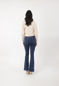 Top de manga larga beige claro con patrón texturizado, combinado con jeans acampanados azul oscuro y zapatos de tacón alto crema. Vista trasera.