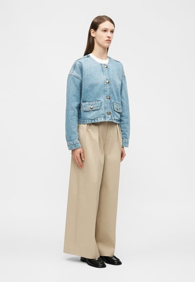 Γυναίκα που φοράει ανοιχτό μπλε denim μπουφάν με κουμπιά, μπεζ παντελόνι φαρδιάς γραμμής και μαύρα loafers, στέκεται μπροστά σε απλό ανοιχτό φόντο.