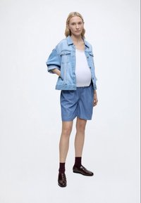 Femeie însărcinată purtând o jachetă și pantaloni scurți din denim albastru deschis, bluză albă, șosete închise la culoare și pantofi maro, stând cu o mână în buzunar.