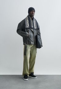 Μαύρο δερμάτινο μπουφάν με φερμουάρ, ελιάς πράσινο παντελόνι φαρδιάς γραμμής, μαύρα αθλητικά παπούτσια και σκούφος σκούρου χρώματος, ολοκληρωμένο με ένα μαντήλι με σχέδια.