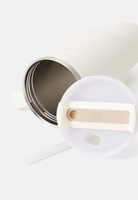 Bouteille en acier inoxydable isolée de couleur crème avec un couvercle blanc, dotée d'une poignée beige et d'un petit orifice pour boire.