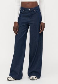 Jeans de denim de pierna ancha en azul oscuro, con una cintura alta, bolsillos clásicos y una ligera textura. Combinados con una camiseta de manga larga blanca y zapatillas.
