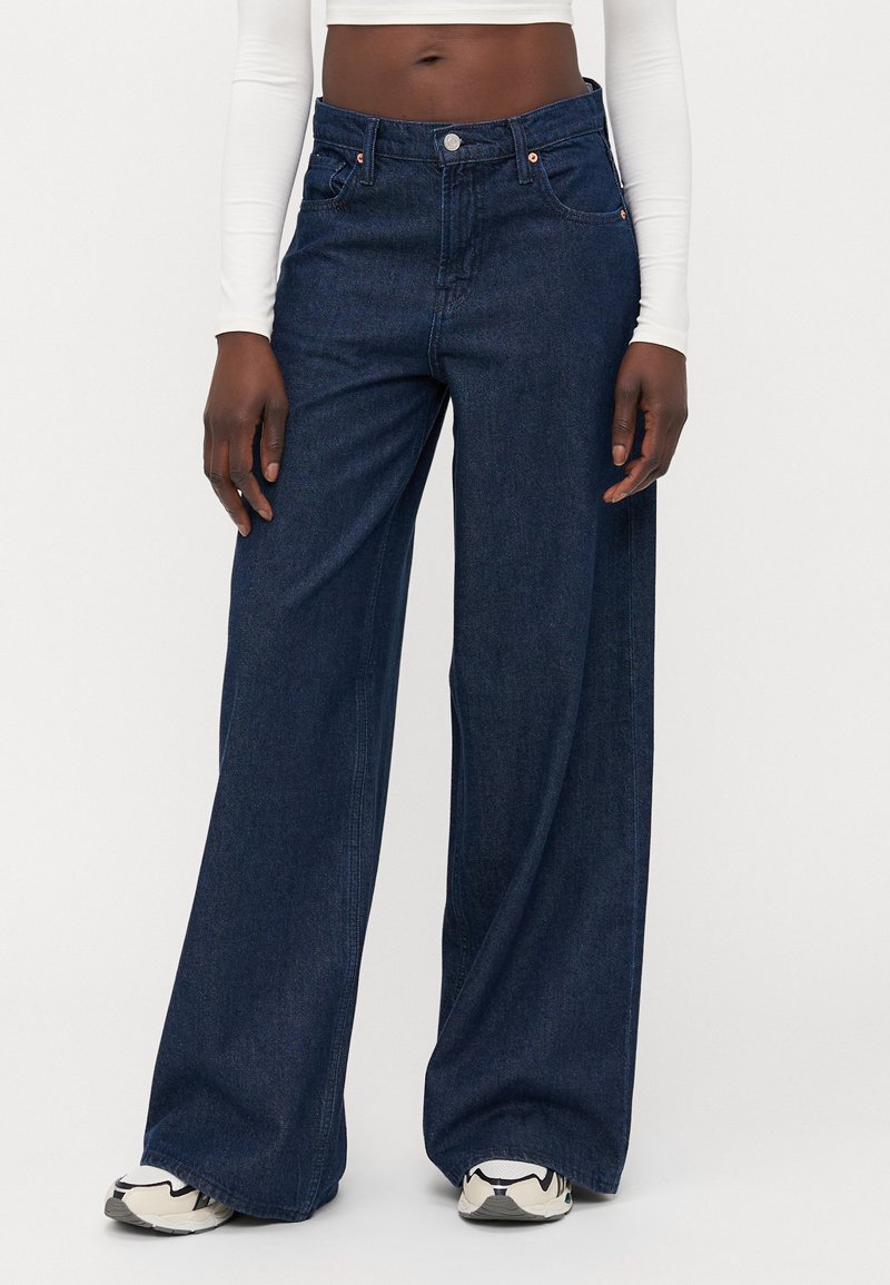 Jeans de denim de pierna ancha en azul oscuro, con una cintura alta, bolsillos clásicos y una ligera textura. Combinados con una camiseta de manga larga blanca y zapatillas.