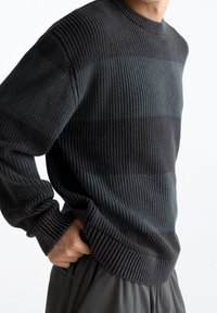 Homme portant un pull en tricot côtelé gris foncé avec de subtiles bandes horizontales de couleur, main dans la poche, sur fond blanc uni.
