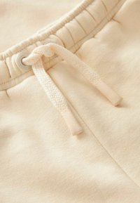 Pantalons de survêtement beige clair avec une finition texturée, dotés d'un cordon de serrage avec des embouts en plastique et des œillets en métal. Tissu doux et décontracté visible.