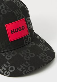 Casquette noire en tissu texturé, motif en relief "HUGO", et un patch en cuir rouge vif avec "HUGO" imprimé en noir.