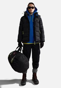 Giubbotto puffa nero con dettagli verde neon sopra un felpa blu, pantaloni cargo neri e scarponi neri. Porta una grande borsa a tracolla nera con toppe.