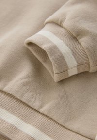 Manche de pull beige en maille avec une bande blanche près du poignet, montrant une texture détaillée et des bords de tissu côtelés.