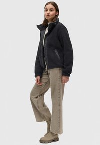 Schwarze Jacke mit Fleece-Futter, glattem Frontpanel, elastischen Bündchen und Seitentaschen, kombiniert mit hellbeigen, geraden Hosen und beigen Stiefeln.