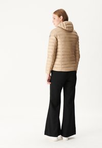 Beige pufferjack met horizontale stiksels en een capuchon, gecombineerd met zwarte flared broek en witte schoeisel, gezien vanuit de achterkant.
