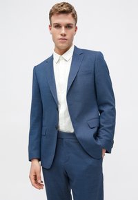 Costum albastru realizat la comandă dintr-un material texturat, cu un design cu un singur rând de nasturi și două butoane, asortat cu o cămașă albă cu nasturi.