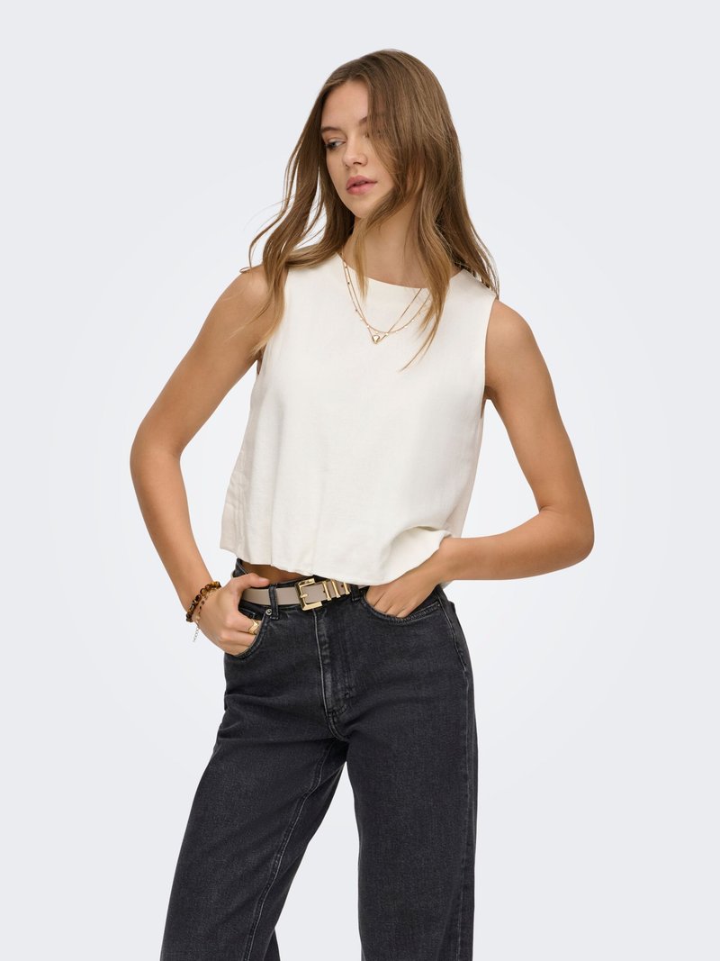 Giovane donna che indossa una canottiera bianca, jeans neri a vita alta con cintura, collana d'oro e braccialetto, posando con le mani nelle tasche.