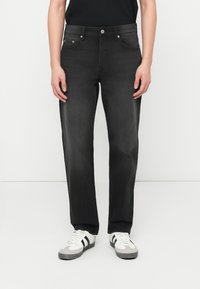 Άτομο με μαύρο τζιν παντελόνι ίσιας γραμμής, μαύρο αμάνικο τοπ και λευκά αθλητικά παπούτσια με μαύρες ρίγες, που στέκεται μπροστά σε απλό λευκό φόντο.