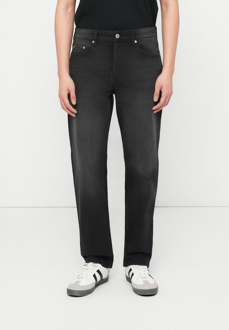 Άτομο με μαύρο τζιν παντελόνι ίσιας γραμμής, μαύρο αμάνικο τοπ και λευκά αθλητικά παπούτσια με μαύρες ρίγες, που στέκεται μπροστά σε απλό λευκό φόντο.