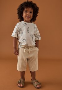 Šviesiai smėlio spalvos marškinėliai su juodomis saulės, palmių ir banglentės raštais, derinami su smėlio spalvos šortais ir alyvinės žalumos sandalais su sagties detalėmis.