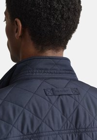 Tmavě modrá prošívaná bunda s vysokým límcem. Vyznačuje se vzorem prošívání a štítkem s logem GANT na zádech. Hladká textura a střih na míru.