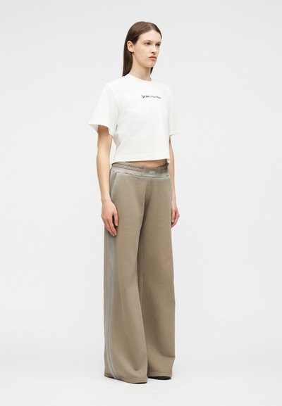 Γυναίκα στέκεται φορώντας λευκό κοντό T-shirt και μπεζ παντελόνι με φαρδιά μούρα και ελαστική μέση με ετικέτα «OFF», πάνω σε μονόχρωμο φόντο.