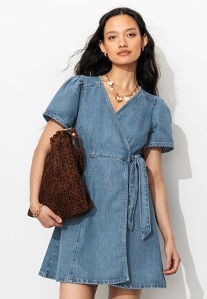 Femme aux longs cheveux foncés portant une robe portefeuille en jean à manches courtes, un collier cœur en or, tenant un sac à imprimé léopard avec un bracelet chaîne en or.