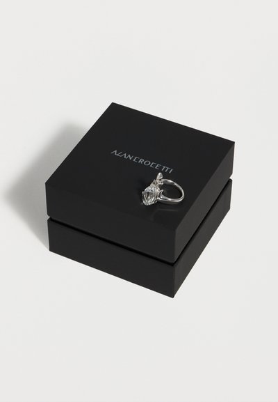 Ασημένιο δαχτυλίδι με μεγάλο διαφανές πολύτιμο λίθο που ακουμπά σε μαύρο κουτί κοσμημάτων Nan Crocetti, σε λευκό φόντο.