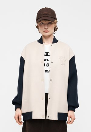 Person wearing beige and navy snap-button varsity jacket, white graphic shirt, brown cap, and hoop earrings, standing against plain background.