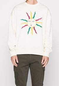 Cremefarbenes Sweatshirt mit einem bunten, bestickten Sonnenmotiv mit geschwungenen Strahlen in Rot, Grün, Gelb und Weiß. Weicher Stoff, lockere Passform.
