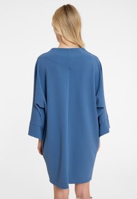 Frau mit blonden Haaren, die ein übergroßes blaues Kleid mit 3/4-Ärmeln trägt, von hinten vor einem schlichten weißen Hintergrund gezeigt.