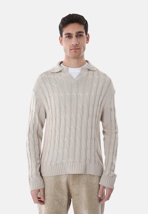 Uomo che indossa un maglione beige a maglia lavorata sopra una camicia bianca e pantaloni marrone chiaro maculati, in piedi davanti a uno sfondo bianco uniforme.
