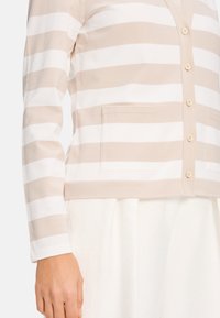 Beige-weißer gestreifter Cardigan mit langen Ärmeln, einem V-Ausschnitt, Vorderknöpfen und zwei aufgesetzten Taschen. Der Stoff wirkt weich und gestrickt.