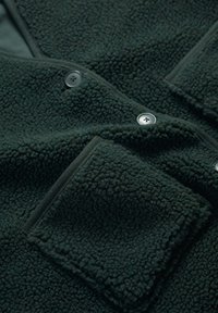 Tamsiai žalias fliso džemperis su tekstūruota paviršiaus, mygtukų uždarymu ir kišene. Minkšta medžiaga, suteikianti jaukumo pojūtį.