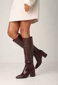 Hnědé kožené vysoké boty s elegantním designem, čtveratou špičkou a masivním podpatkem. Hladká textura s minimálním kovovým zdobením.