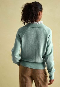 Pull tricoté vert menthe avec un col et des poignets côtelés, présentant un détail de couture le long de la colonne vertébrale. Porté avec un pantalon marron à carreaux.