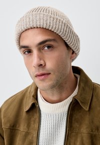 Knit beanie in light beige with ribbed texture, worn with a tan suede jacket and a white textured sweater. Close-up of face.