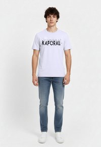 Biała bawełniana koszulka z czarnym napisem "KAPORAL" o różnej grubości, zestawiona z niebieskimi dżinsami i białymi trampkami. Prosty design.