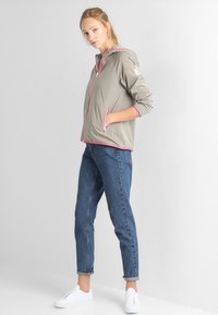 Lichtgrijze jas met roze ritsen en accenten, voorzien van een capuchon en twee zijzakken, gecombineerd met blauwe spijkerbroek en witte sneakers.