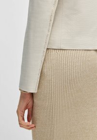 Lichtbeige blazer met rafelige randen in combinatie met een geribbelde, metallic gouden rok. De texturen contrasteren, wat een gestroomlijnde silhoeët benadrukt.