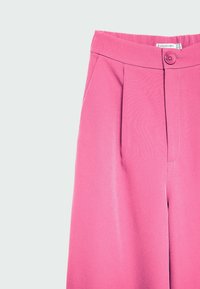 Höga rosa byxor med knäppning, framfickor och en synlig varumärkesetikett inuti midjebandet.