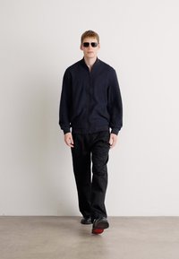 Casual outfit met een donkerblauwe bomberjack met rits, zwarte broek en donkere zonnebril, gecombineerd met stijlvolle schoenen en een minimalistisch ontwerp.